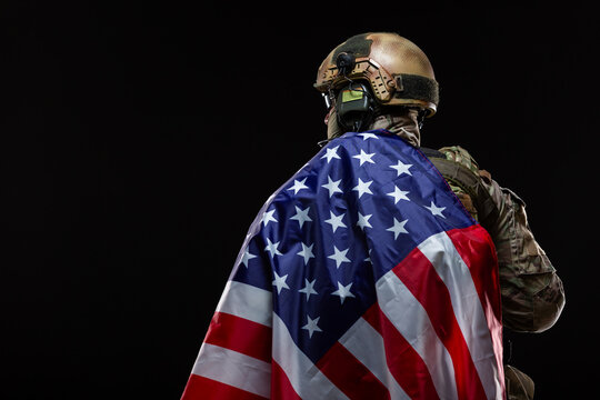 Back view of a military man in a hard hat and full gear, draped over the shoulder of flag of America isolated on black. The concept of patriotism, protection of the Motherland, love for the country