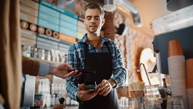Tall Caucasian Customer Pays for Coffee and Pastry with Contactless NFC Payment Technology on Smartphone to a Handsome Barista in Blue Checkered Shirt. Contactless Mobile Payment in Cafe Concept.