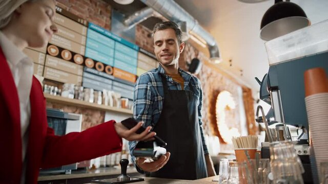 Female Customer Pays for Take Away Coffee with Contactless NFC Payment Technology on Smartphone to a Handsome Barista in Checkered Shirt in Cafe. Customer Uses Mobile to Pay Through Bank Terminal.