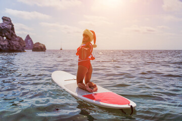 Happy young attractive brunette woman in red swimsuit and Santa hat, swimming on sup board in the...