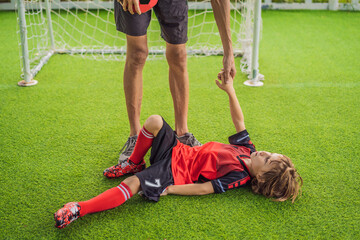 Fototapeta premium Little cute kid boy in red football uniform fell on the football field and his trainer or father playing soccer, football on field, outdoors. Active child making sports with kids or father, Smiling