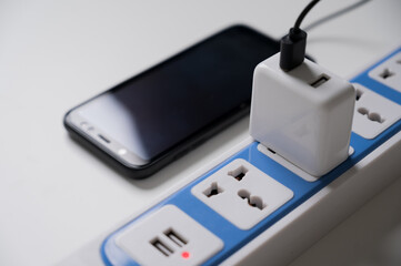 The smartphone is charged on a power strip on the white table. Electrical equipment, electrical wires and power strips in the house.
