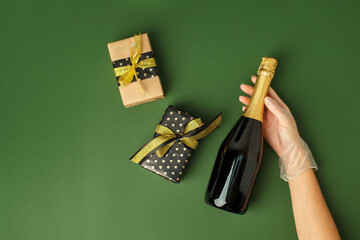 Medical latex Gloved hands hold a bottle of champagne and gifts on natural green background Party concept, winter holiday in terms of pandmias, coronavirus, self-isolation, banner, copy space