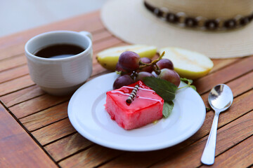 Still life of food. White plate with red cake and grapes on a wooden table. Next to a plate a cup of black coffee, a spoon and a sun hat. The concept of rest and travel.