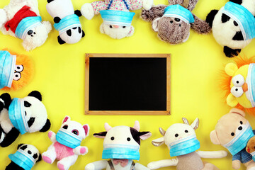 Many toys in protective face masks. Chalkboard surrounded by stuffed toys on yellow background. Top view. Flat lay, copy space. Coronavirus protection concept.