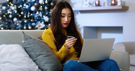 Happy excited Asian young woman sitting in decorated room on Christmas and buying xmas presents online on laptop paying with credit card for purchase on internet. Holiday shopping. Discounts concept