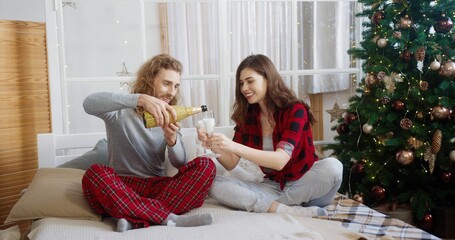 Handsome Caucasian young man sitting in room near decorated christmas tree with beloved woman and popping bottle of shampagne celebrating New Year together at home. Family celebration. Xmas concept