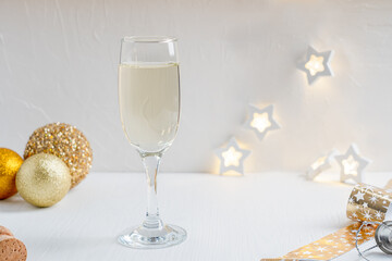 Wineglass full of dry white Prosecco sparkling champagne with golden glittering christmas or new year decoration ball served on white wooden table against garland defocus lights. Horizontal image