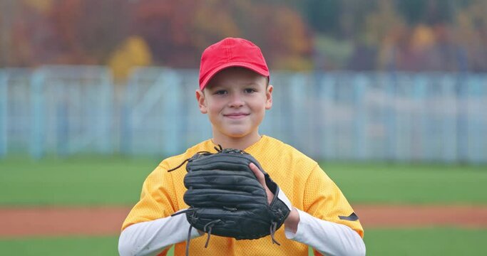 Portrait of a boy baseball player on a blurry background, the pitcher holds the ball in his glove and looks at the camera, smiling boy, 4k slow motion.