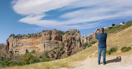 Fototapeta na wymiar A male tourist photographs the famous arched bridge of Ronda in Andalusia in summer with a cloudy blue sky. The city is on top of the plateau. At the very bottom is a small waterfall.