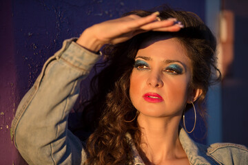 A beautiful young woman in a denim jacket with bright makeup in the style of the eighties stands...