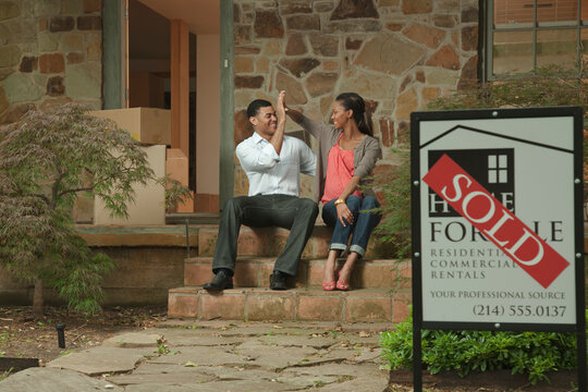 Couple Sitting On Front Stoop Of House With Sold Sign