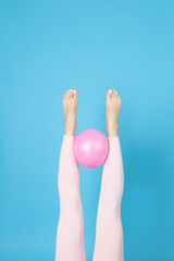 women's legs in pink sports leggings hold a rubber Pilates ball between them, isolated on a blue background.
