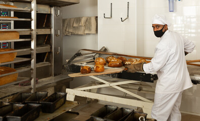 Baker of small bakery in face mask for viral protection holding hot bread on shovel just from oven