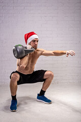 Obraz premium Athletic man in a Santa hat squats with a kettlebell.