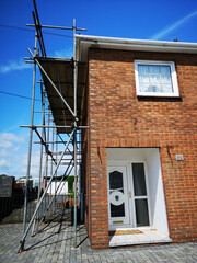Traditional red brick detached house undergoing renovation or repairs with scaffolding and boards attached to the house. Low angle, corner view with blue sky.