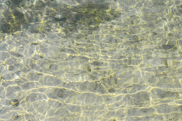 Fototapeta na wymiar Shallow clear green and yellow water with algae and small stones on the bottom. Shiny light reflections pattern and waves