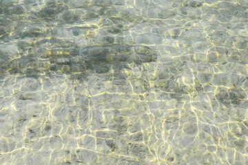 Shallow clear green and yellow water with algae and small stones on the bottom. Shiny light reflections pattern and waves