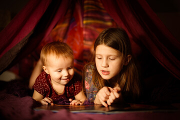 Caucasian girls children sisters read a book in a toy tent with garlands, children's games and houses, the older sister reads to the younger, dark cozy photos,