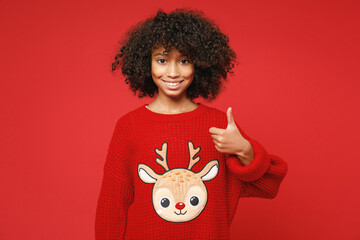 Little african american curly kid girl 12-13 years old in knitted cozy deer Christmas sweater showing thumb up isolated on bright red background children studio portrait Childhood lifestyle concept.
