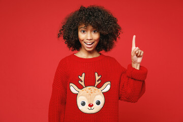 Little african american curly kid girl 12-13 years old wearing knitted deer Christmas sweater finger up idea isolated on bright red background children studio portrait Childhood lifestyle concept