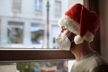 Ragazzo con cappello da babbo natale e mascherina ffp2 guarda triste fuori casa attraverso il vetro...