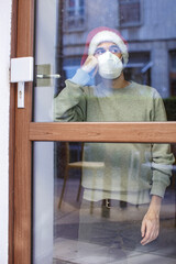 Uomo con cappello da babbo natale e mascherina ffp2 guarda triste fuori casa attraverso il vetro del portoncino 