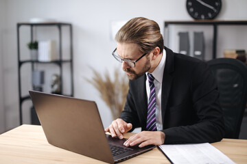 Concentrated bearded man dressed in stylish suit working on wireless laptop while sitting at bright office. Handsome businessman in eyewear using modern gadget for work.