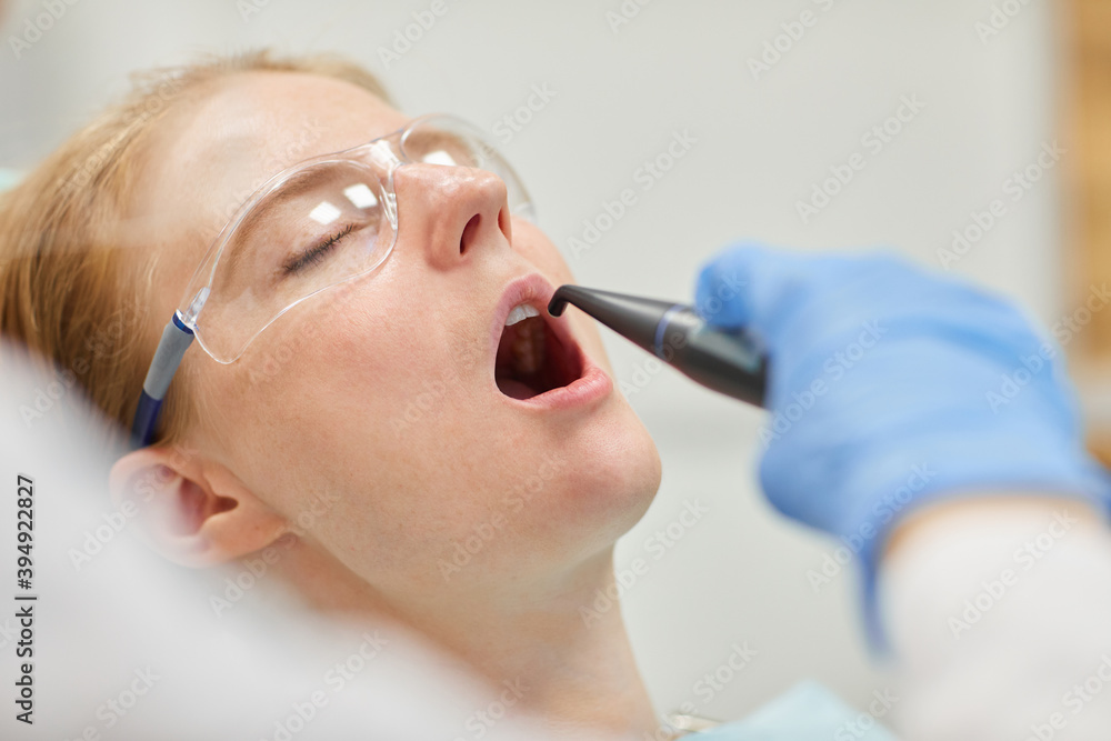 Wall mural Female patient in protective glasses lying with eyes closed and opened mouth while dentist treating her teeth