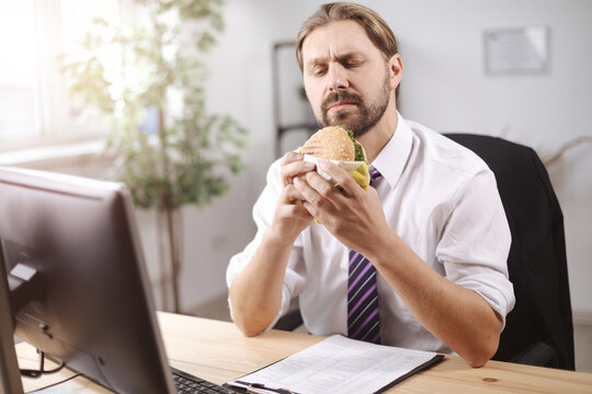 Mature man in white shirt and black tie eating junk food during working time at office. Bearded businessman having no time for healthy lunch at work.
