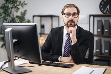Front view of confident bearded man in black suit and eyeglasses sitting at desk with modern computer. Mature businessman enjoying favorite work at office center.