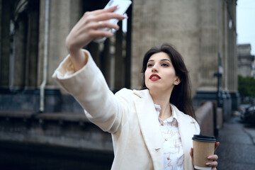 Beautiful model woman  making selfie on the background of attractions. The girl makes a selfie with coffee on the street.