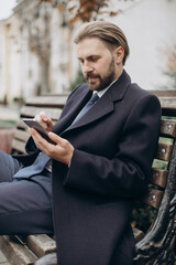 Happy bearded man relaxing on wooden bench outdoors and using modern smartphone. Mature businessman browsing internet during leisure time on fresh air.
