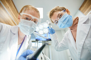 Portrait of two dentists in protective masks and glasses looking at camera while working with medical instruments in team