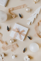 Paper gift box with bow tie and Christmas toys: fir-trees, baubles, ribbons on pastel beige background. Holiday celebration concept. Top view, flat lay. Christmas / New Year decoration.