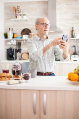 Mature man surfing the social media using phone during breakfast in kitchen. Elderly person using internet online chat technology video webcam making a video call connection camera communication