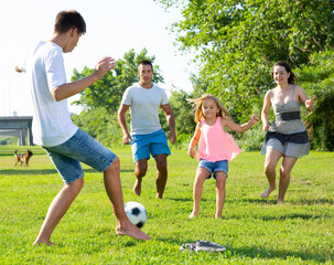 Positive parents with two kids playing soccer together on green field on summer day. High quality photo