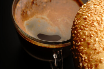 Coffee in a transparent cup with a bagel sprinkled with spices. Close. The light was dim. On a black background.