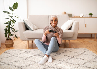 Happy Muslim woman in hijab sitting on floor with joystick, playing videogame at home, full length...