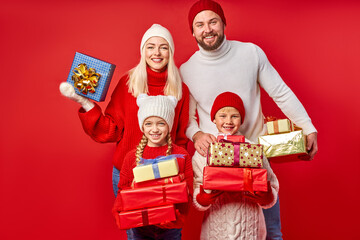 portrait of open-minded positive family with gifts boxes isolated on red background, studio shoot. young parents celebrate New Year with children, look at camera