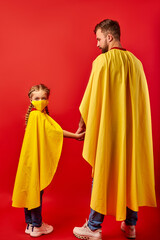 friendly man and kid girl in yellow cloaks, pretend to be superheroes. male and child in mask stand holding hands together, rear view, girl looks at camera