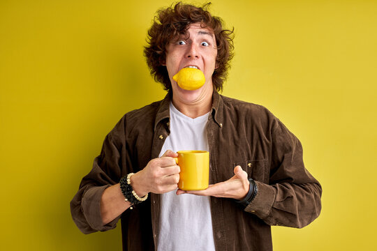 Young Male Stuffed His Mouth With Lemon, Want To Be Healthy. Hold Cup In Hands Isolated Over Yellow Green Background