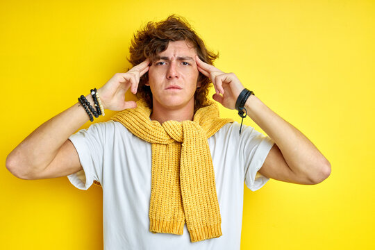 thoughtful male keeps hand on temple isolated on yellow wall, suffers from headache, remembers yesterday party. students, youth and lifestyle concept