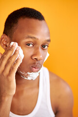 young adult male is applying shaving foam before shaving bristle in the morning, studio shoot of guy with dark skin caring for skin, isolated orange background