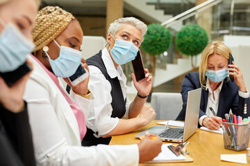 team of diverse business women in medical masks coworking in office during pandemic, coronavirus concept. ladies sit at table talking, in formal wear