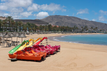 Fototapeta premium Colorful water slides boats docked on the empty white sand beach unused because of lack of tourism and lockdown measures at Playa el Camison, Las Americas, Tenerife, Canary Islands, Spain