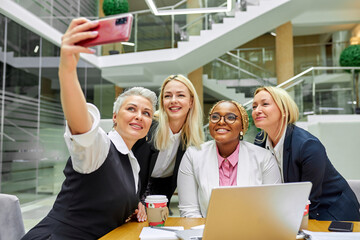 portrait of diverse business women taking selfie on smartphone, make selfie, laugh, friendly ladies in light modern office