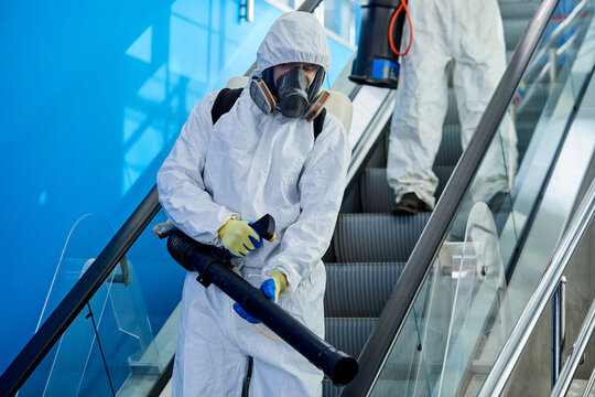 male disinfector cleaning the area in building, disinfect escalator, wearing protective white suit, getting rid of coronavirus infections around