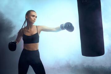 fit brunette woman in boxing gloves and sportswear kicking bag on dark background with smoke.