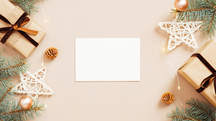 Christmas flat lay composition with empty paper card mockup, gift boxes, stars decoration, fir tree branches on beige background. Cozy home table, hygge style.